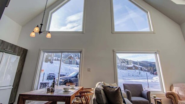 A high-ceilinged room with large windows, a dining table, and seating.