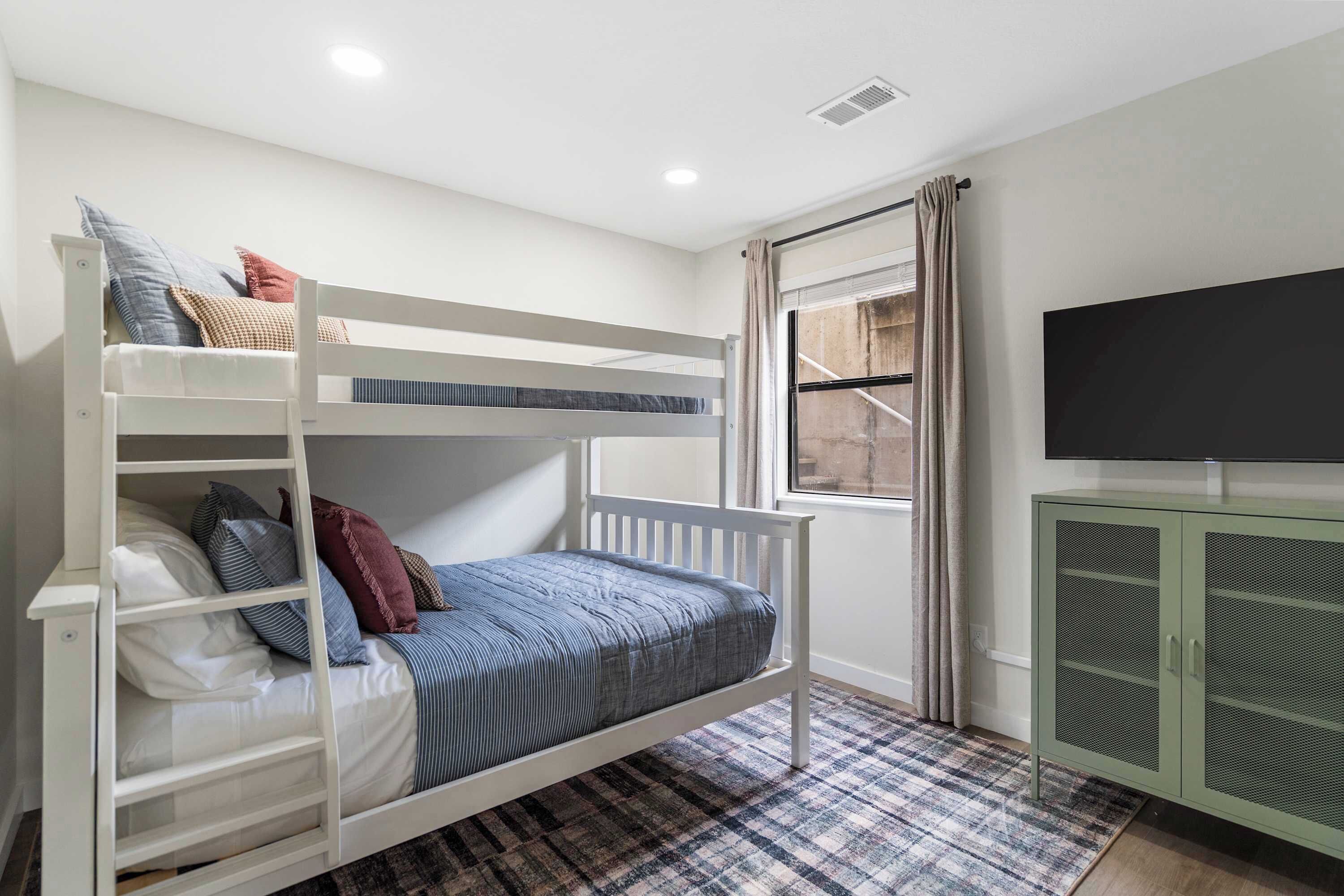 A bunk bed with pillows and a blanket, a television on a cabinet, and a window with curtains.