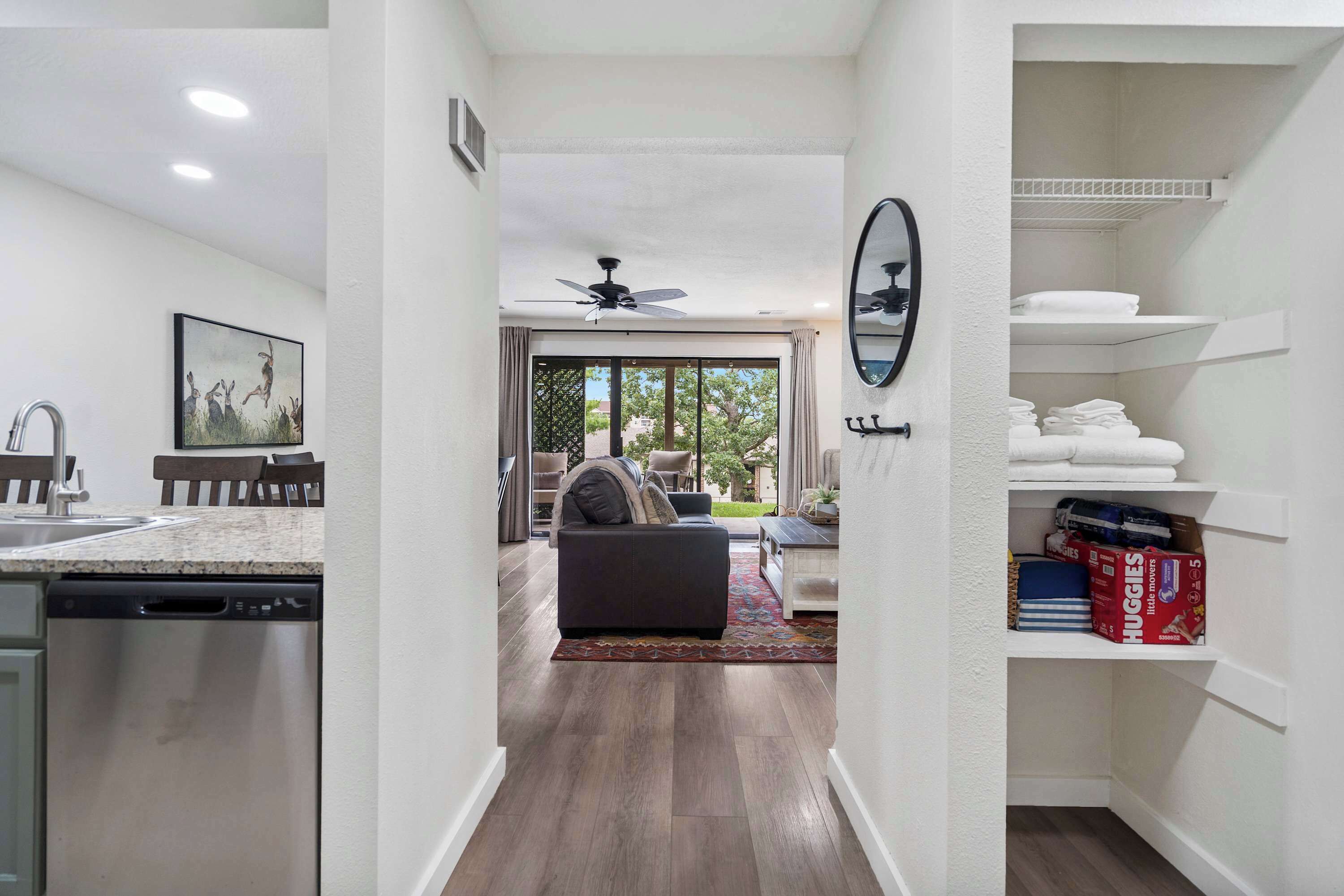 A modern kitchen with a dishwasher, a dining area with a sofa and chairs, a ceiling fan, and a view through a glass door to an outdoor area.