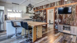 A modern kitchen with a wooden island, bar stools, a large wooden dining table, and a TV mounted on a wooden wall.