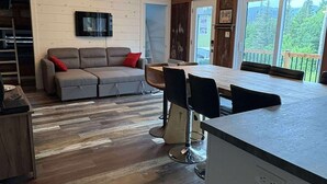 A modern kitchen with a central island, a dining area with chairs, a sofa with cushions, and a TV mounted on the wall.