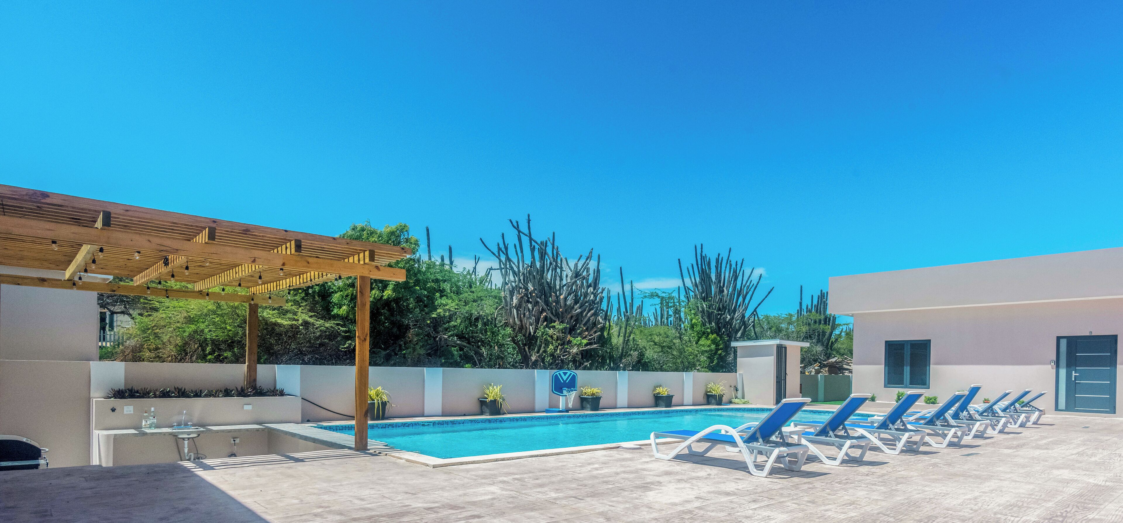 A pool area with lounge chairs, a pergola, and a small building.