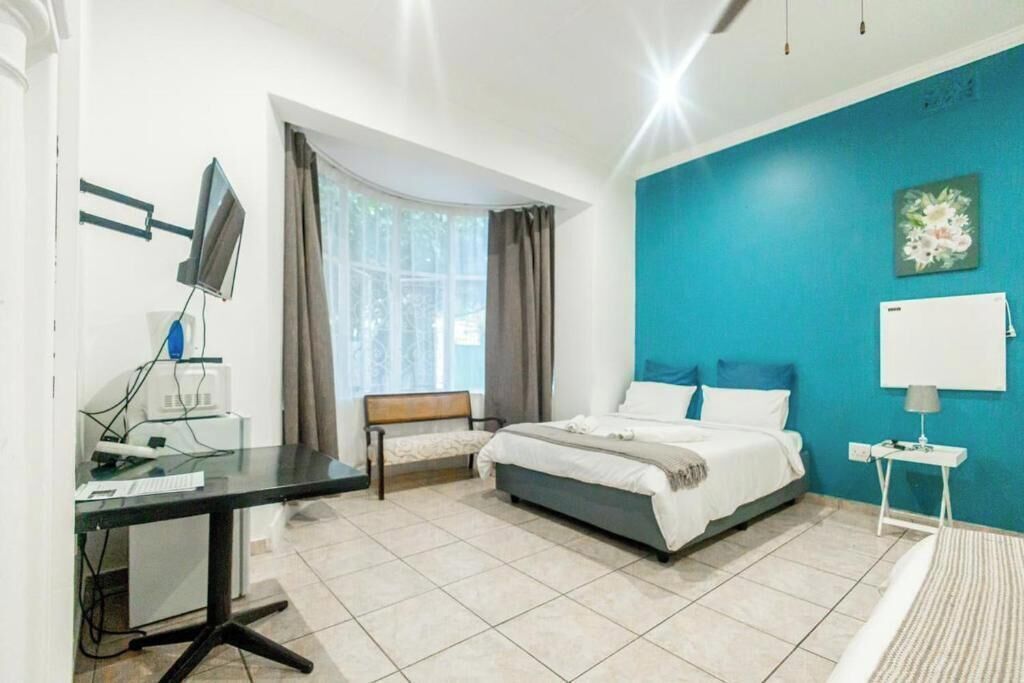A hotel room with a bed, a desk with a computer, a chair, a window with curtains, and a blue accent wall.