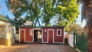 A small, wooden structure with a door and windows, situated in a paved courtyard with trees in the background.