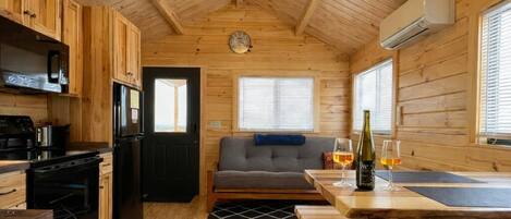 Interior de una cabaña con paredes de madera, una kitchenette, un sofá, una mesa de comedor y un ventilador de techo.