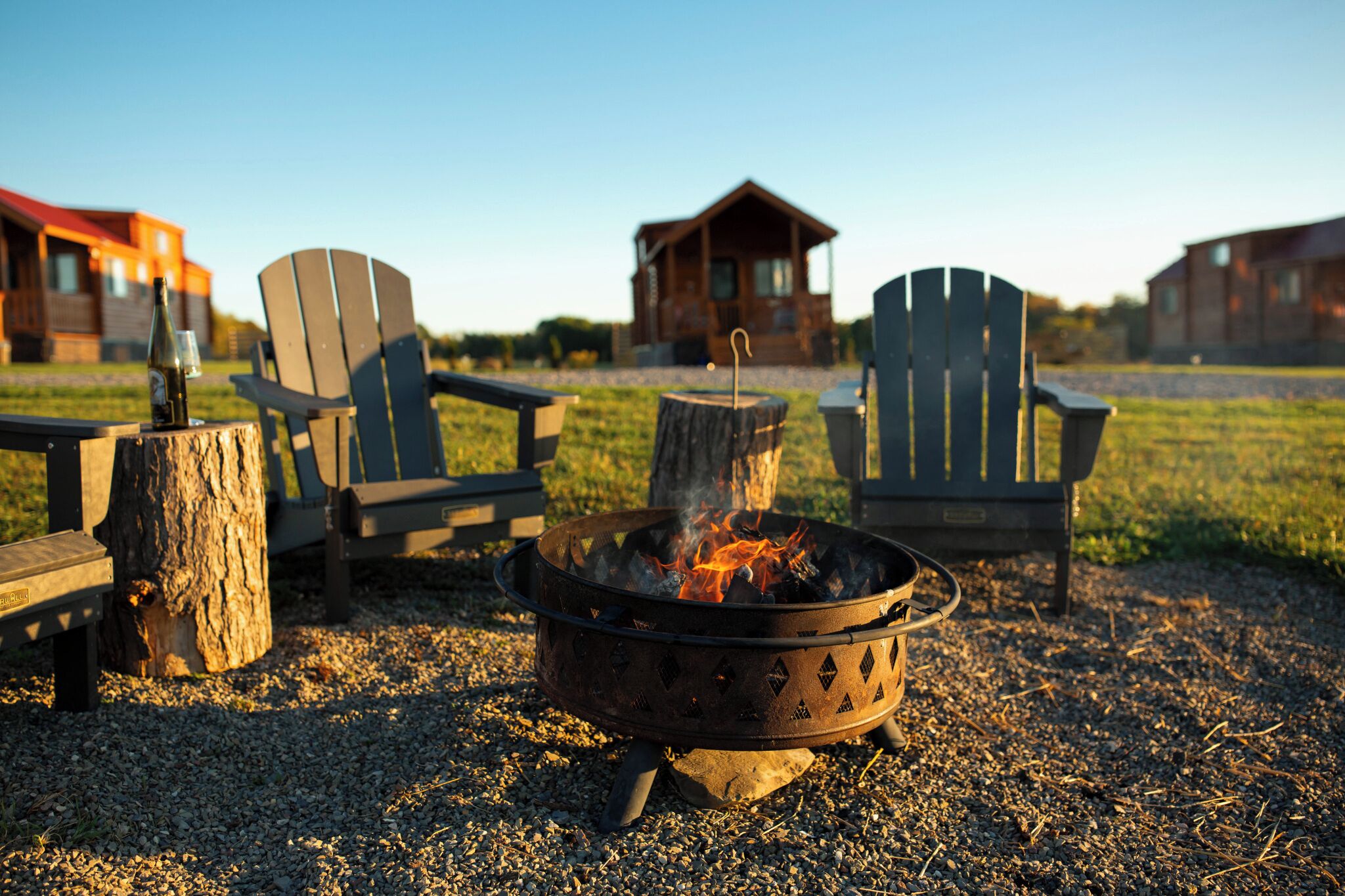 A campfire with two Adirondack chairs, a wooden stump, and a metal fire pit.