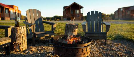 A campfire with two Adirondack chairs, a wooden stump, and a metal fire pit.