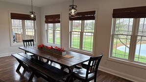 A dining area with a wooden table and benches, large windows with blinds, and hanging pendant lights.