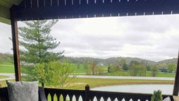 A covered outdoor seating area with wicker furniture and a wooden table, overlooking a green landscape.