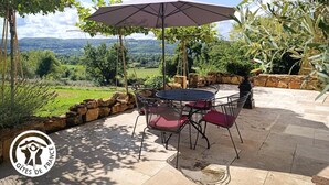 Um pátio externo com mesa e cadeiras, um guarda-sol e vista para uma paisagem verde.