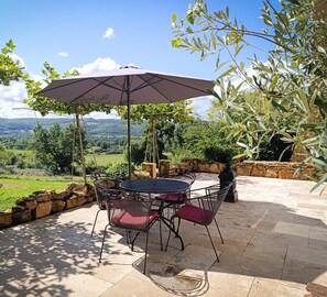 An outdoor patio with a table and chairs, an umbrella, and a view of a green landscape.