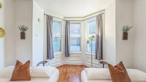 Ein Schlafzimmer im Eckbereich mit zwei Betten, Holzboden und Blick auf die Straße durch große, mit Vorhängen versehene Fenster.