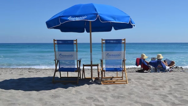 Dos sillas de playa con una sombrilla azul con la leyenda "Oceanside Beach Service" sobre una playa arenosa y el océano al fondo.