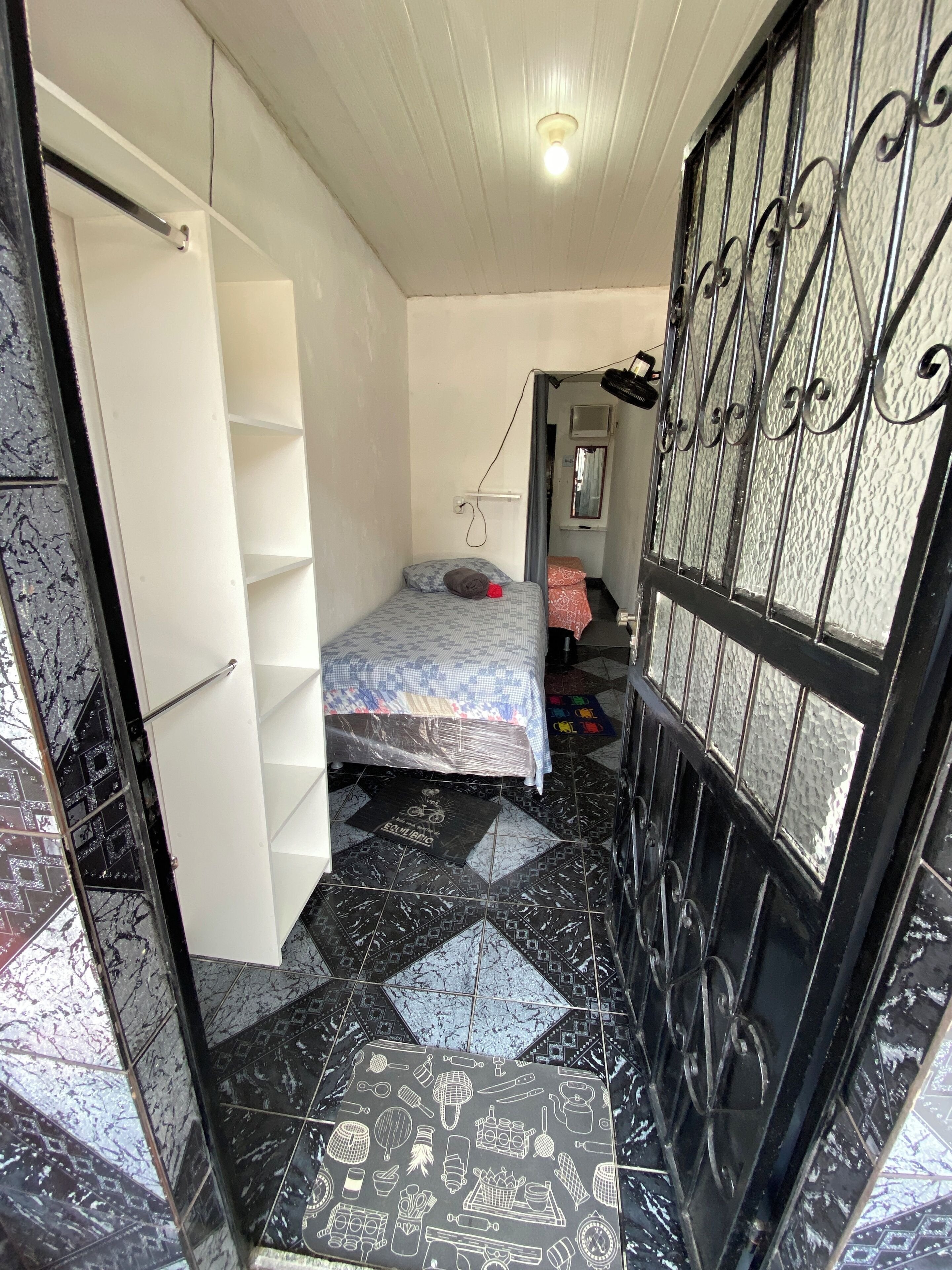Una habitación pequeña y estrecha con una cama, un armario y una puerta que da a otra habitación. El suelo presenta un diseño de baldosas con patrón blanco y negro.