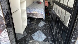 A small, narrow room with a bed, a wardrobe, and a door leading to another room. The floor has a black and white patterned tile design. - Apartment in Coroado, Manaus, close to buses, restaurants and markets. (Manaus)