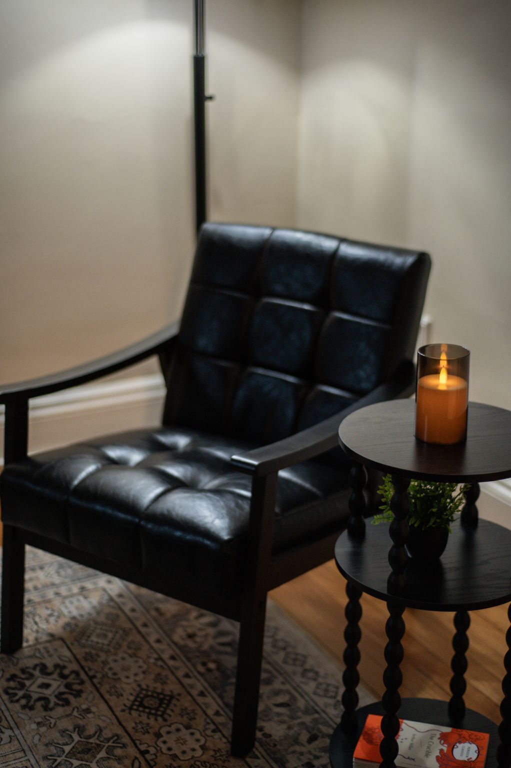 A black leather chair with a cushion, a small round table with a candle, and a patterned rug.