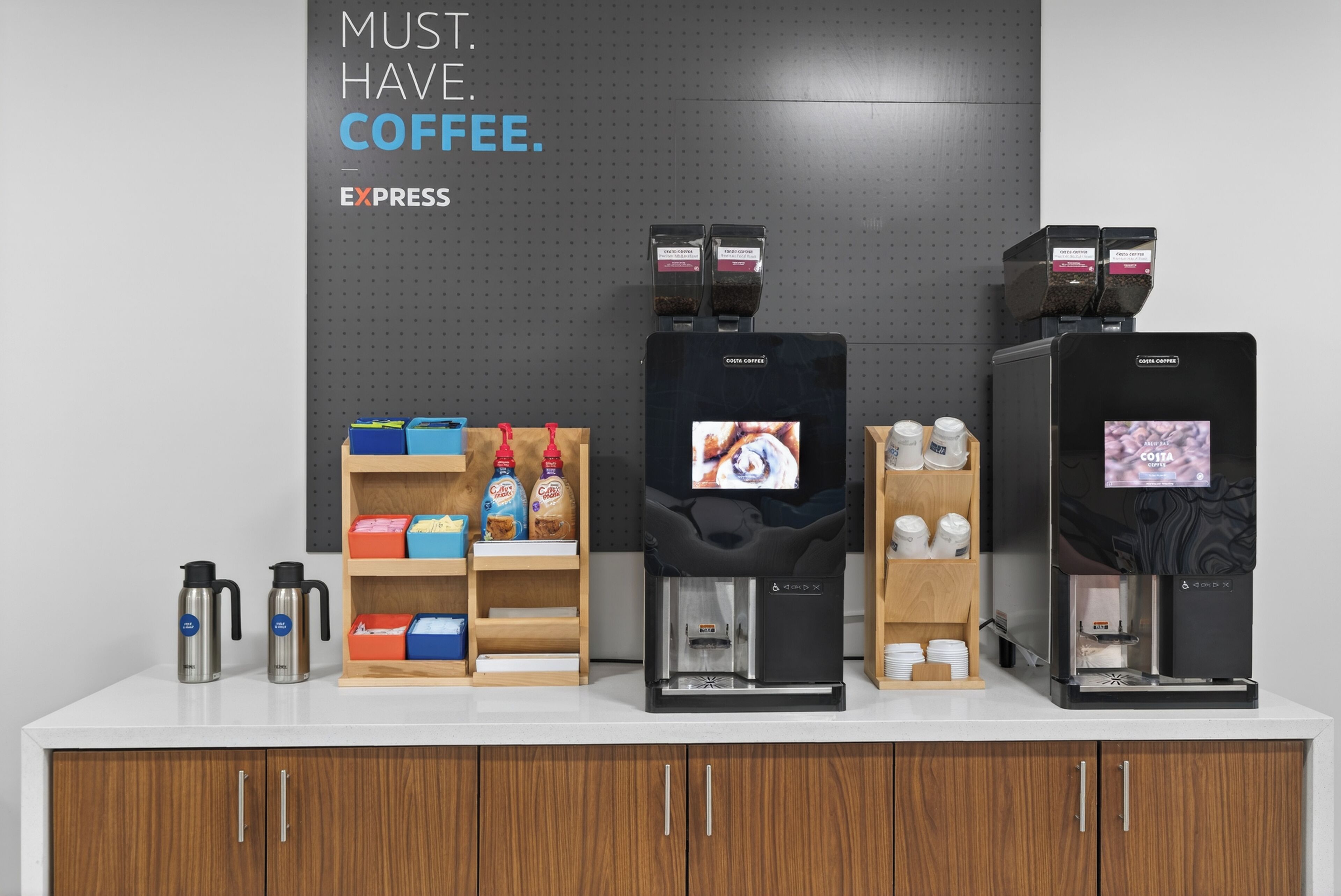 Una estación de café con varios dispensadores, un estante con cápsulas de café y un letrero que dice 'MUST. HAVE. COFFEE'.