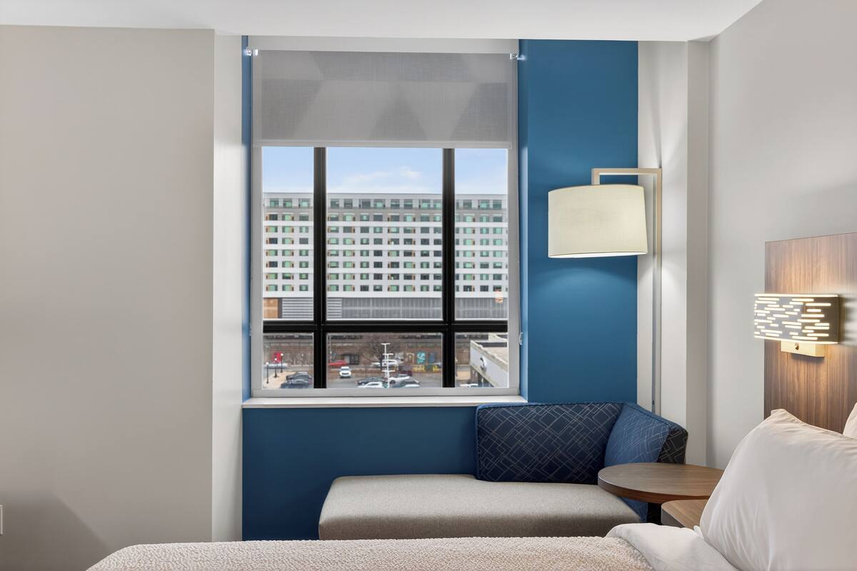 Una habitación de hotel con un ventanal grande, una cama, una mesita pequeña y una lámpara fijada a la pared.