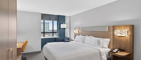 A modern hotel room with a large bed, a desk, and a view of the city through a window.