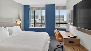 Habitación de hotel con cama, escritorio, silla, televisión y ventanas amplias con vistas a la ciudad.