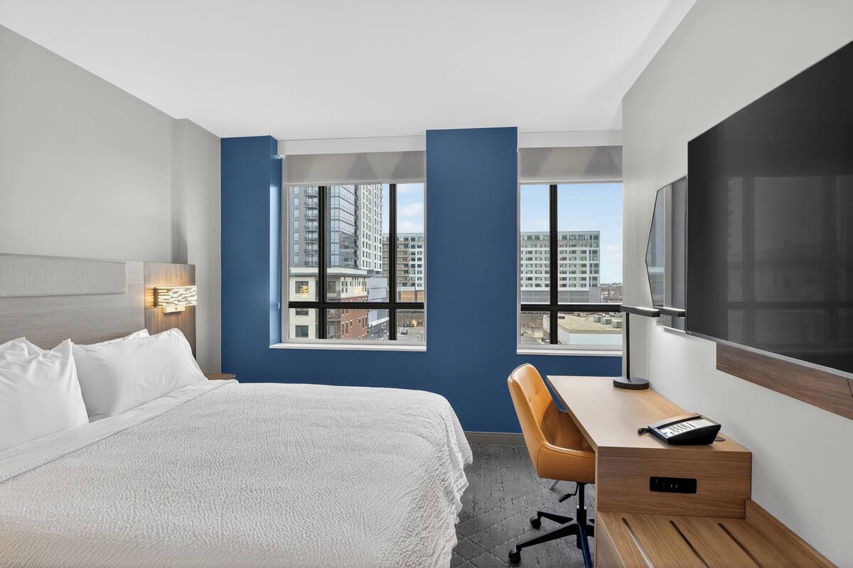 Habitación de hotel con cama, escritorio, silla, televisión y ventanas amplias con vistas a la ciudad.