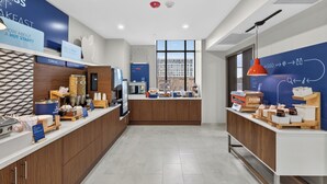 Un moderno comedor con una barra que exhibe cereales y frutas, una máquina de café y vistas de la ciudad a través de amplias ventanas.