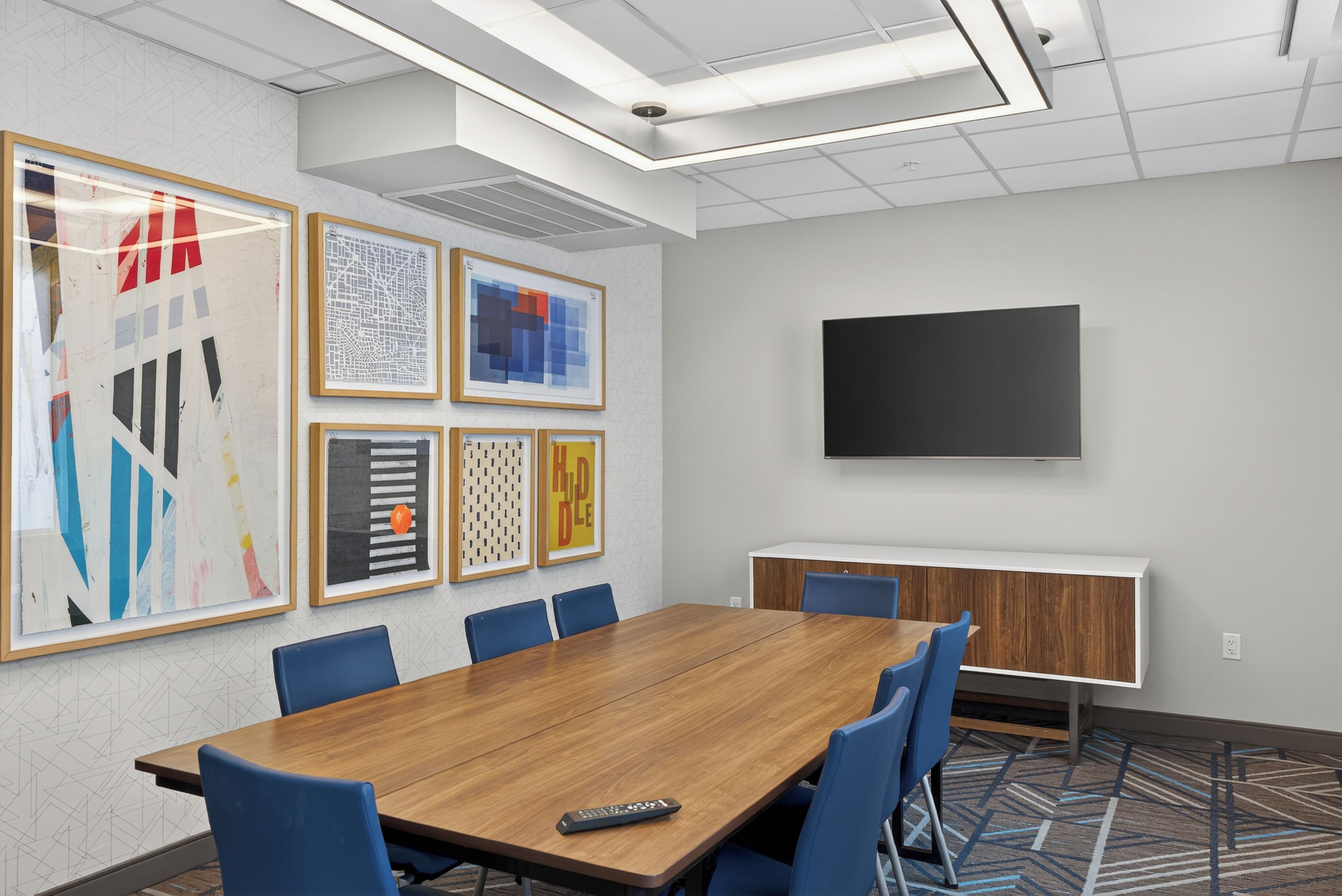 A conference room with a wooden table, blue chairs, a flat-screen TV, and framed artwork on the wall.