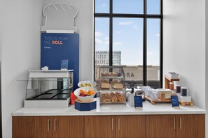 Un buffet de petit-déjeuner proposant une variété de plats, un panneau indiquant les informations relatives aux allergies alimentaires, et une vue sur un bâtiment à travers de grandes fenêtres.