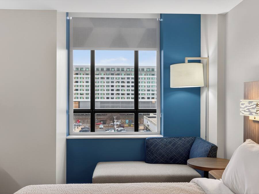 Een hotelkamer met een groot raam, een bed, een klein tafeltje en een wandlamp.