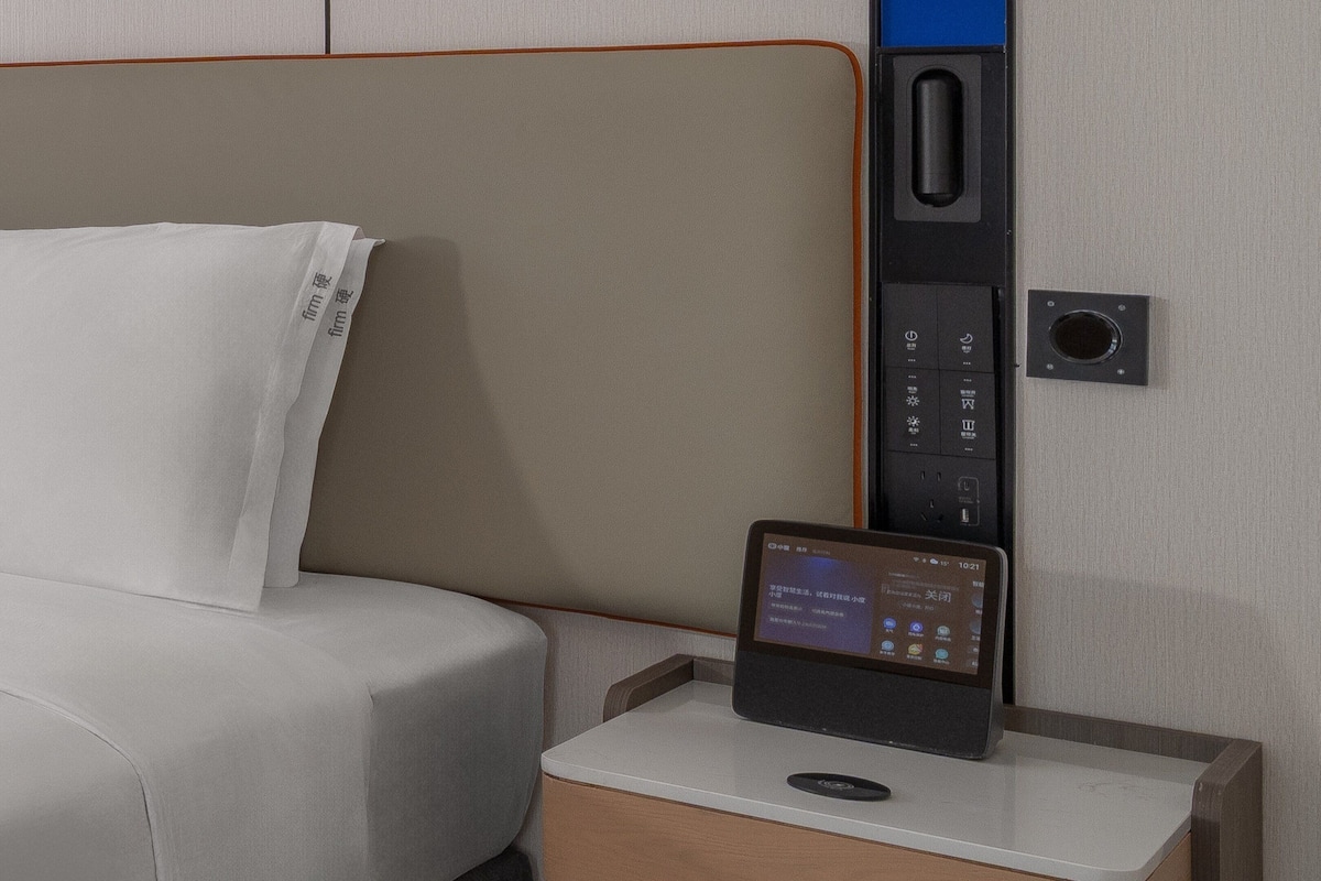 Ein modernes Hotelzimmer mit einem ordentlich bezogenen Bett, einem Nachttisch mit einem Tablet, das Informationen zum Zimmer anzeigt, und einer Wandbedienung.