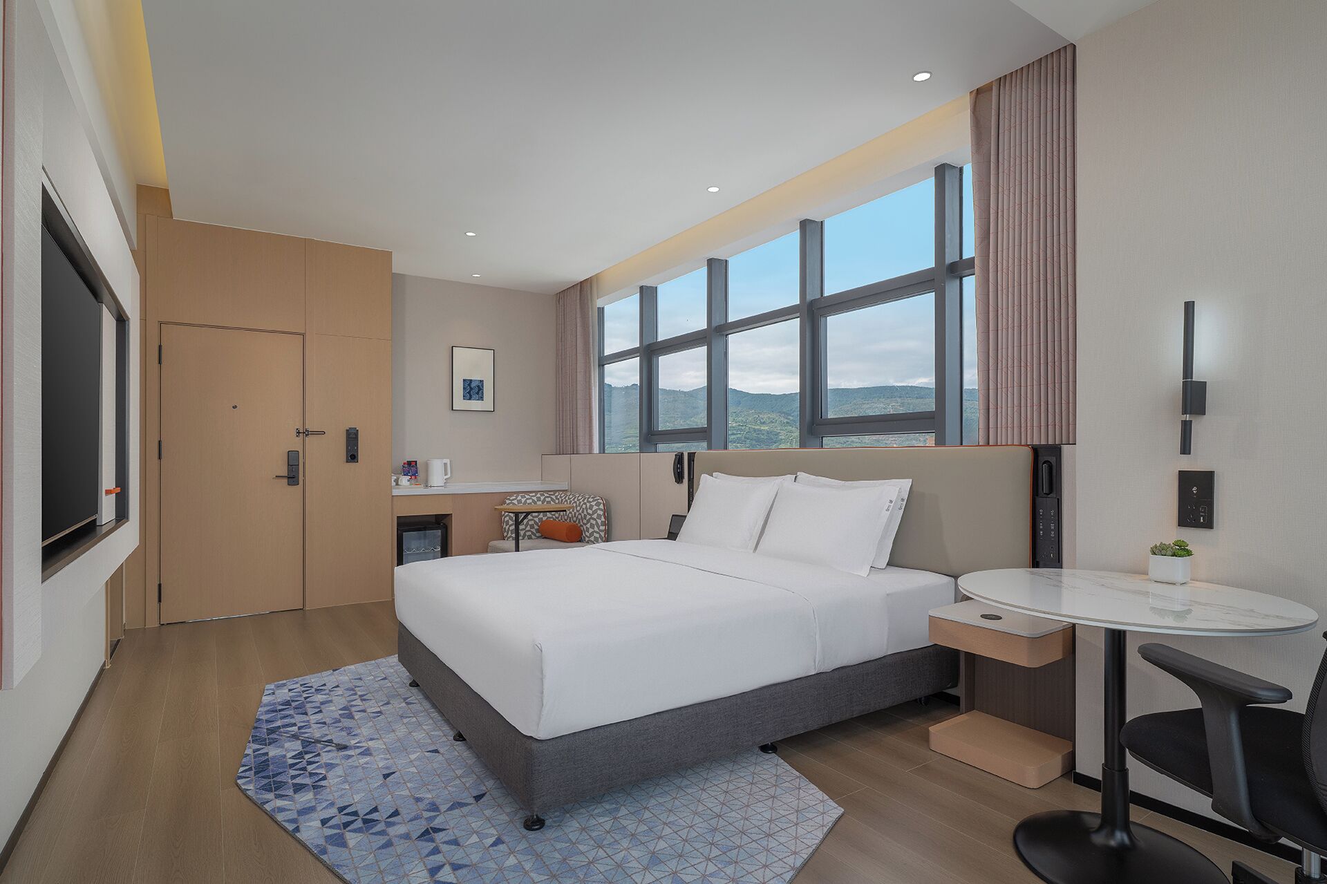 A modern hotel room with a large bed, a small desk, a chair, a window with a view, and a door.