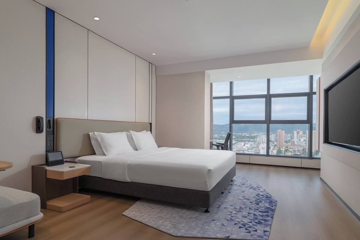 Ein modernes Hotelzimmer mit einem großen Bett, einem Schreibtisch mit Stuhl, einem Flachbildfernseher und Blick auf die Stadt durch große Fenster.