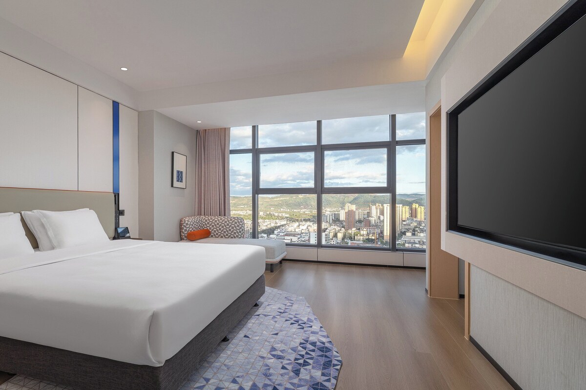 Een moderne hotelkamer met een groot bed, een zithoek en uitzicht op de stad door grote ramen.