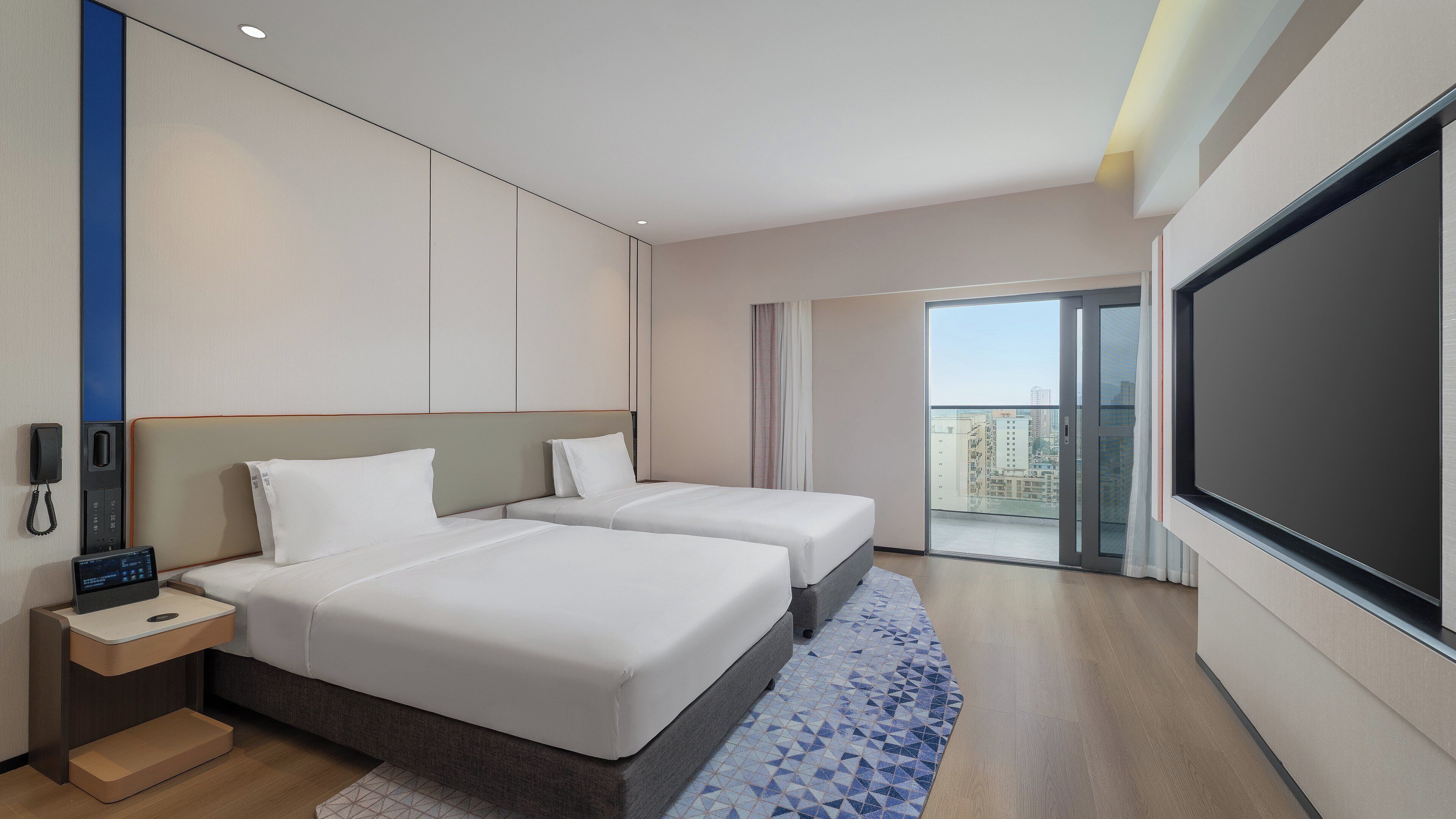 Ein Hotelzimmer mit zwei Betten, einem Schreibtisch mit Telefon, einem Fernseher und einem Balkon mit Blick auf die Stadt.