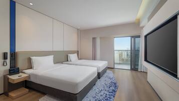 Ein Hotelzimmer mit zwei Betten, einem Schreibtisch mit Telefon, einem Fernseher und einem Balkon mit Blick auf die Stadt.
