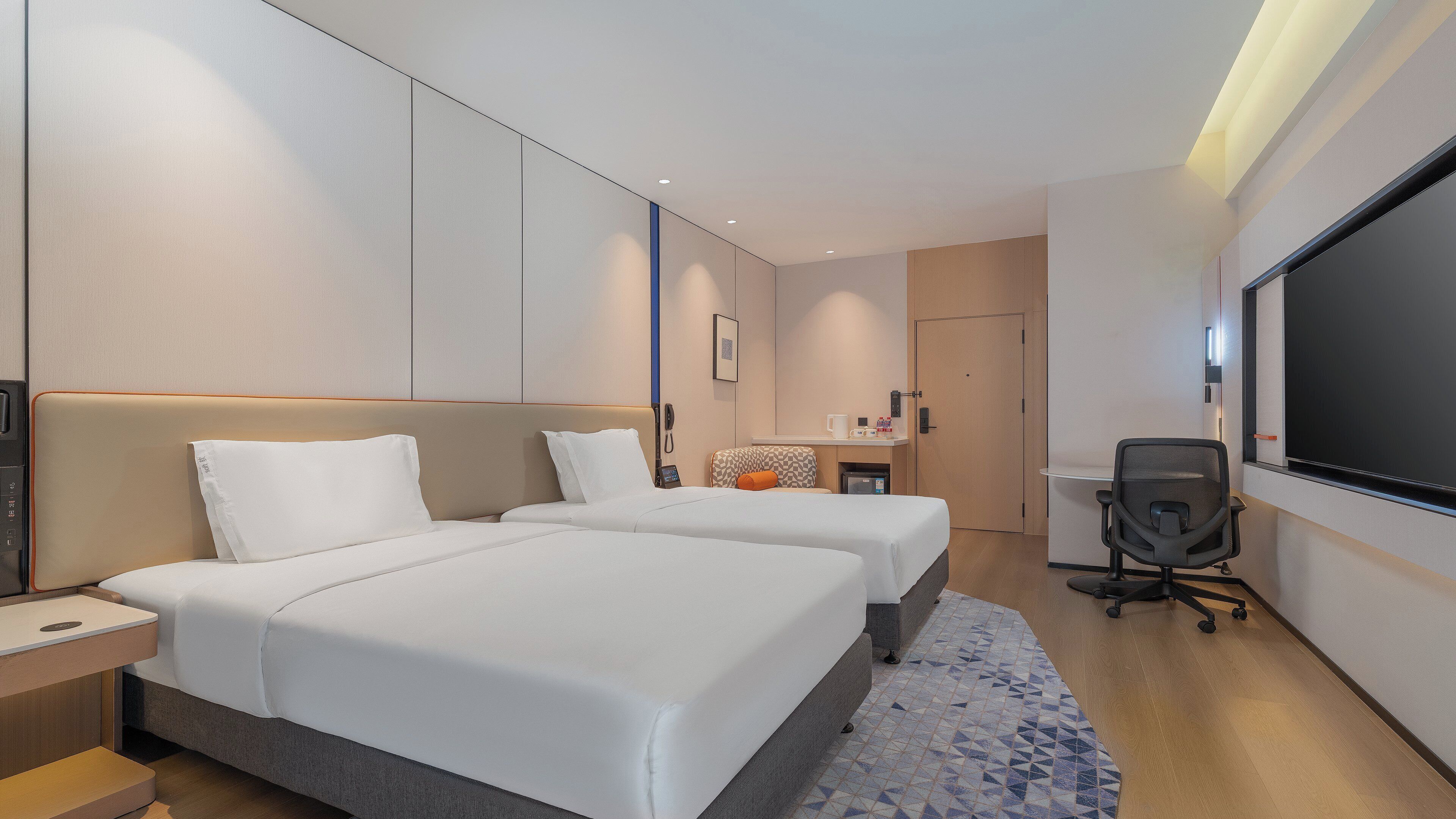 A hotel room with two beds, a desk, a chair, a TV, and a small kitchenette.