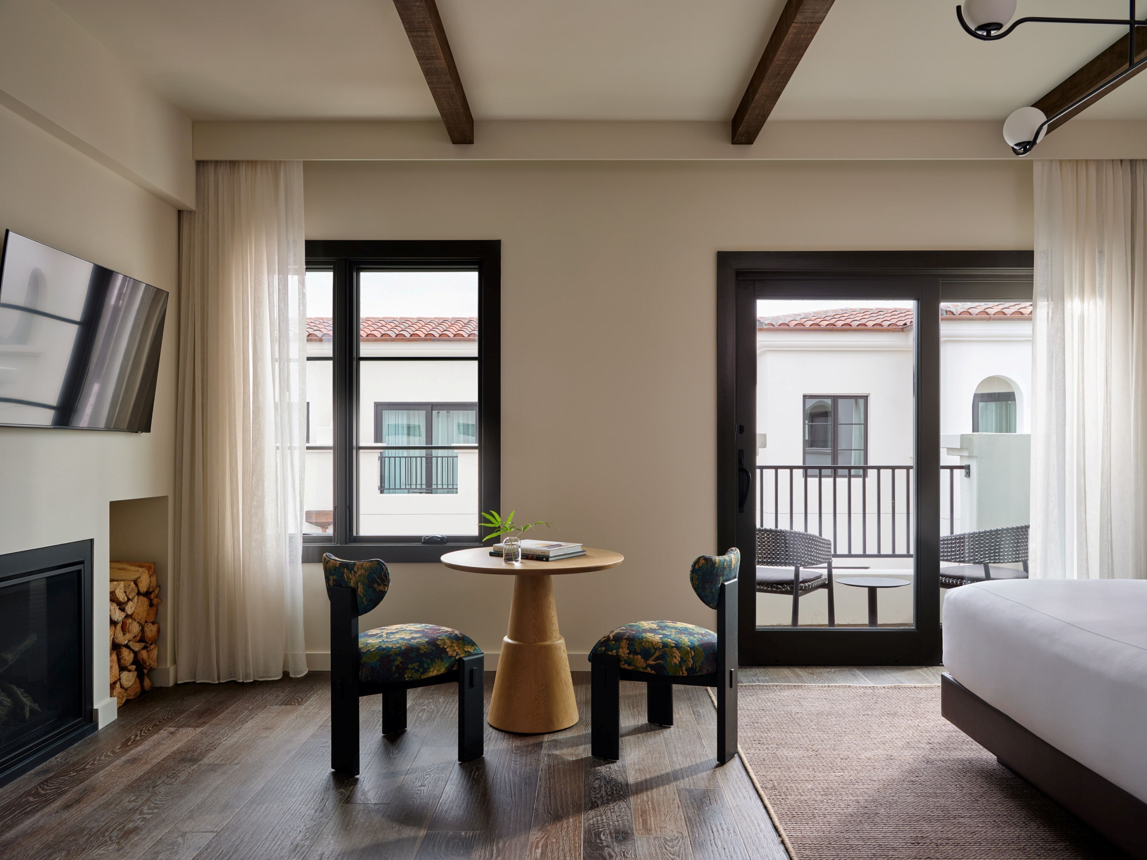 A hotel room with a bed, a TV, a fireplace, a balcony with a view, and two chairs with a small table.