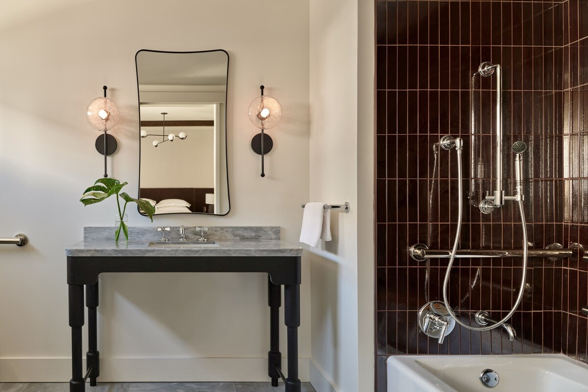 A modern bathroom with a marble vanity, two wall-mounted lights, a large mirror, a potted plant, a towel rack, and a separate shower area with a glass enclosure.