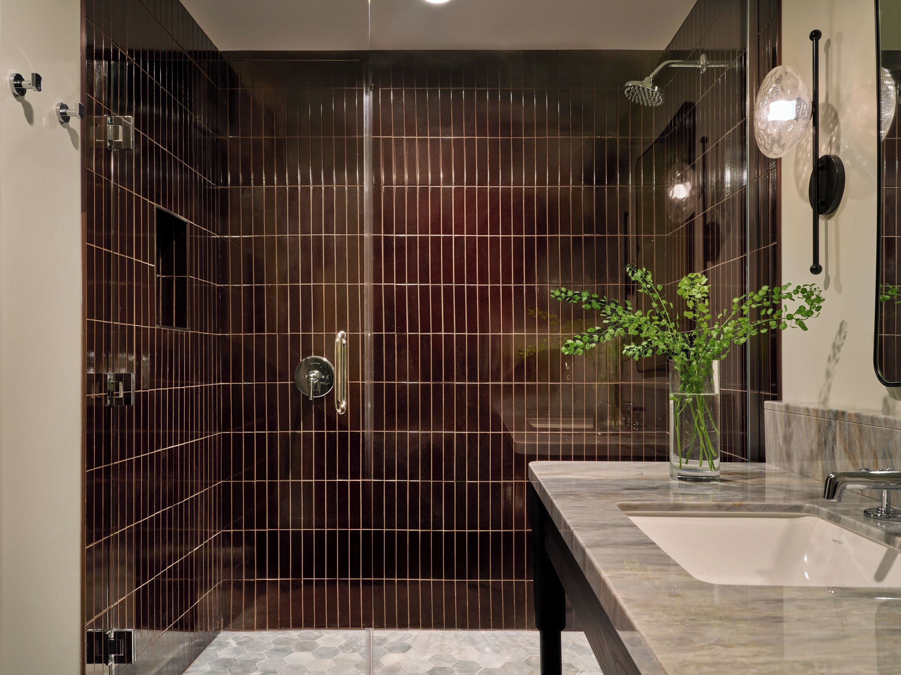 A modern bathroom with a dark tiled shower wall, a glass shower enclosure, a marble sink, and a potted plant.
