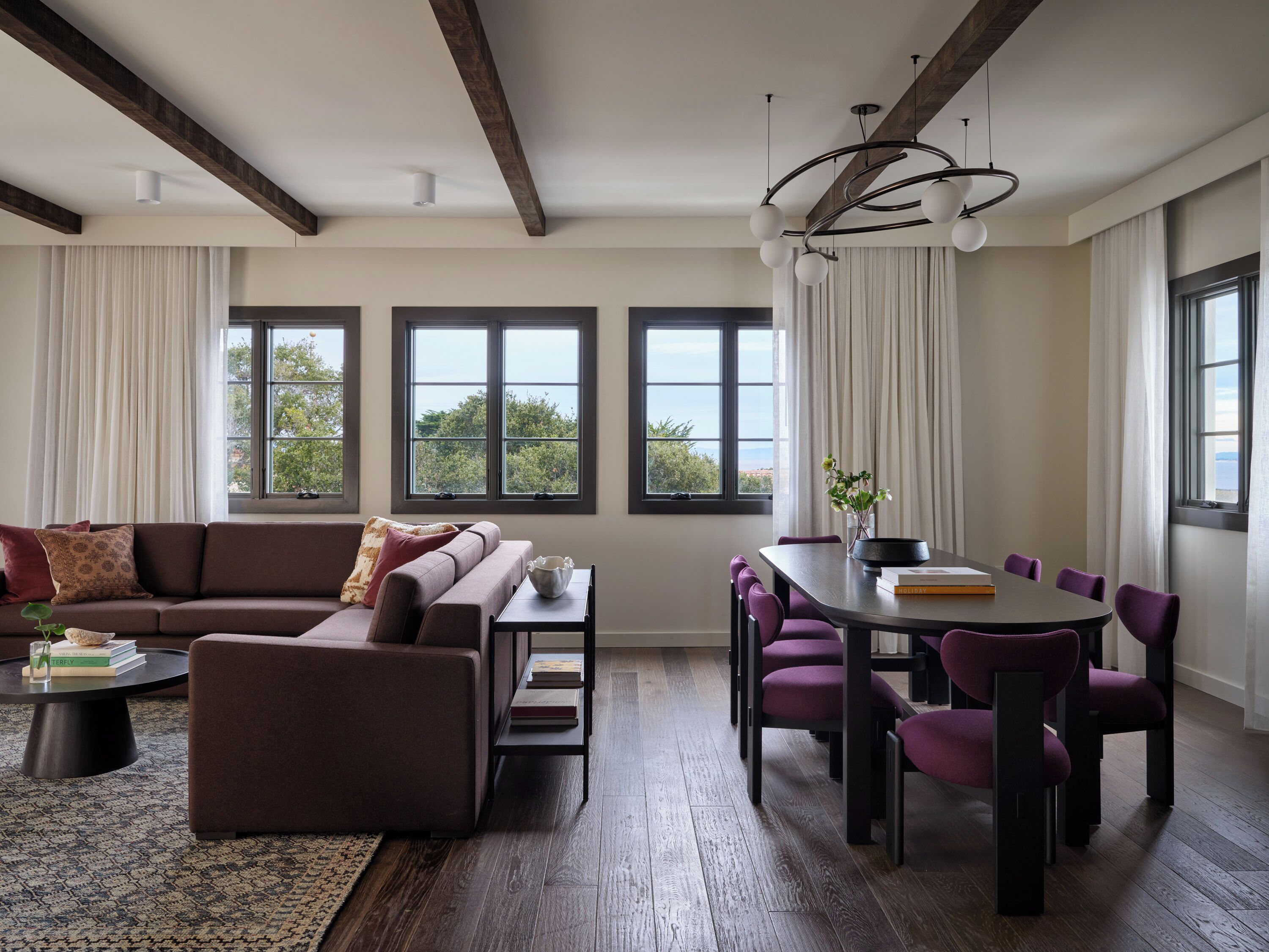 Un salon moderne avec un canapé marron, une table basse et un coin repas avec des chaises violettes. De grandes fenêtres sont ornées de rideaux, un lustre illumine la pièce et l’on peut admirer la verdure à l’extérieur.