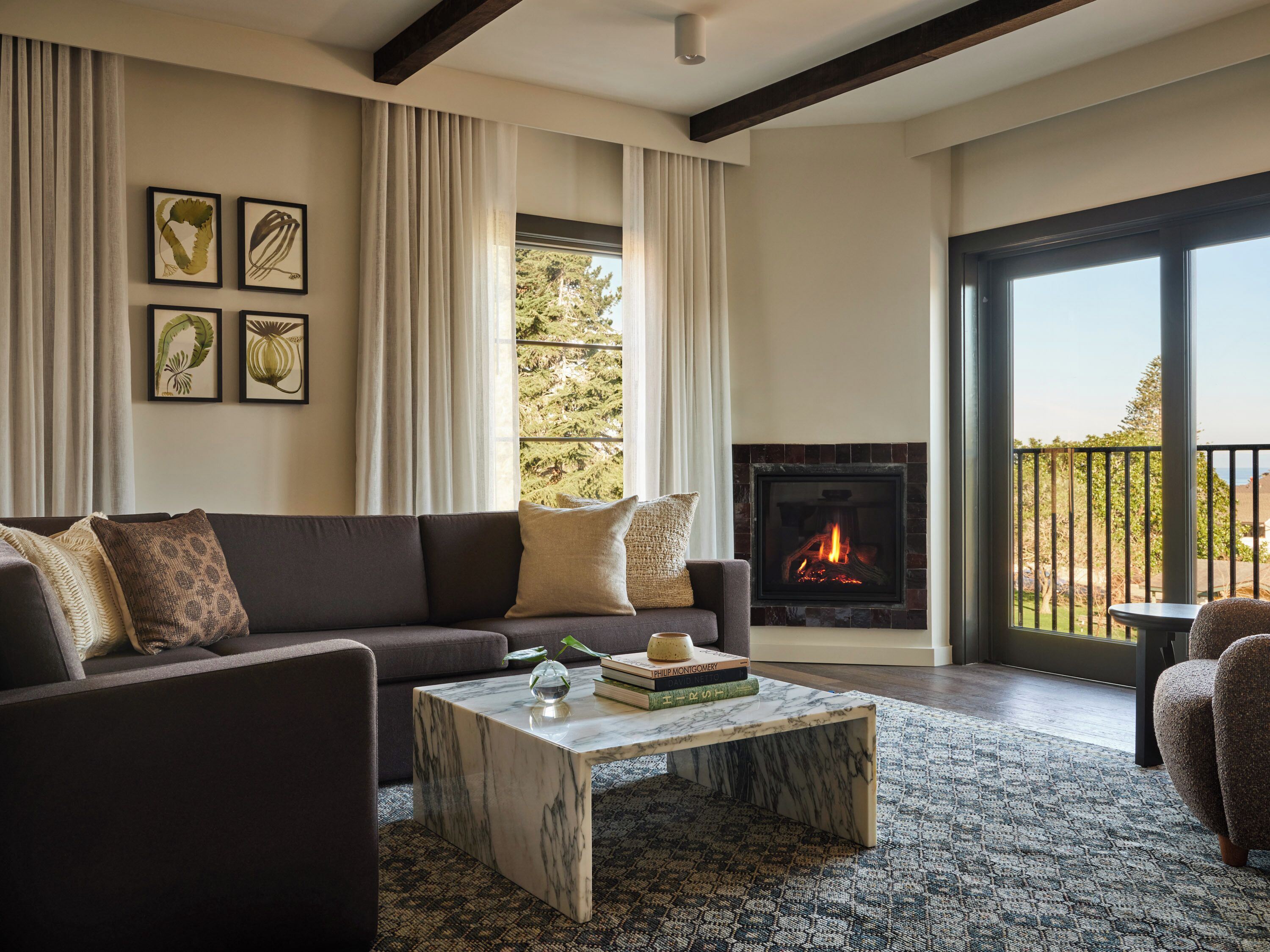 A modern living room with a fireplace, a sofa, a coffee table, and a view of trees through large windows.