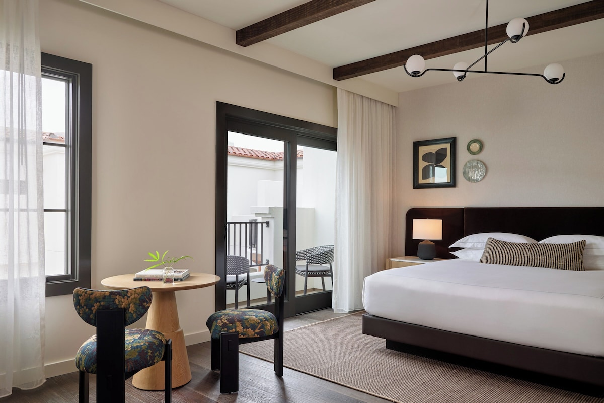 A hotel room with a large bed, a round table with two chairs, a balcony door, and a ceiling light fixture.