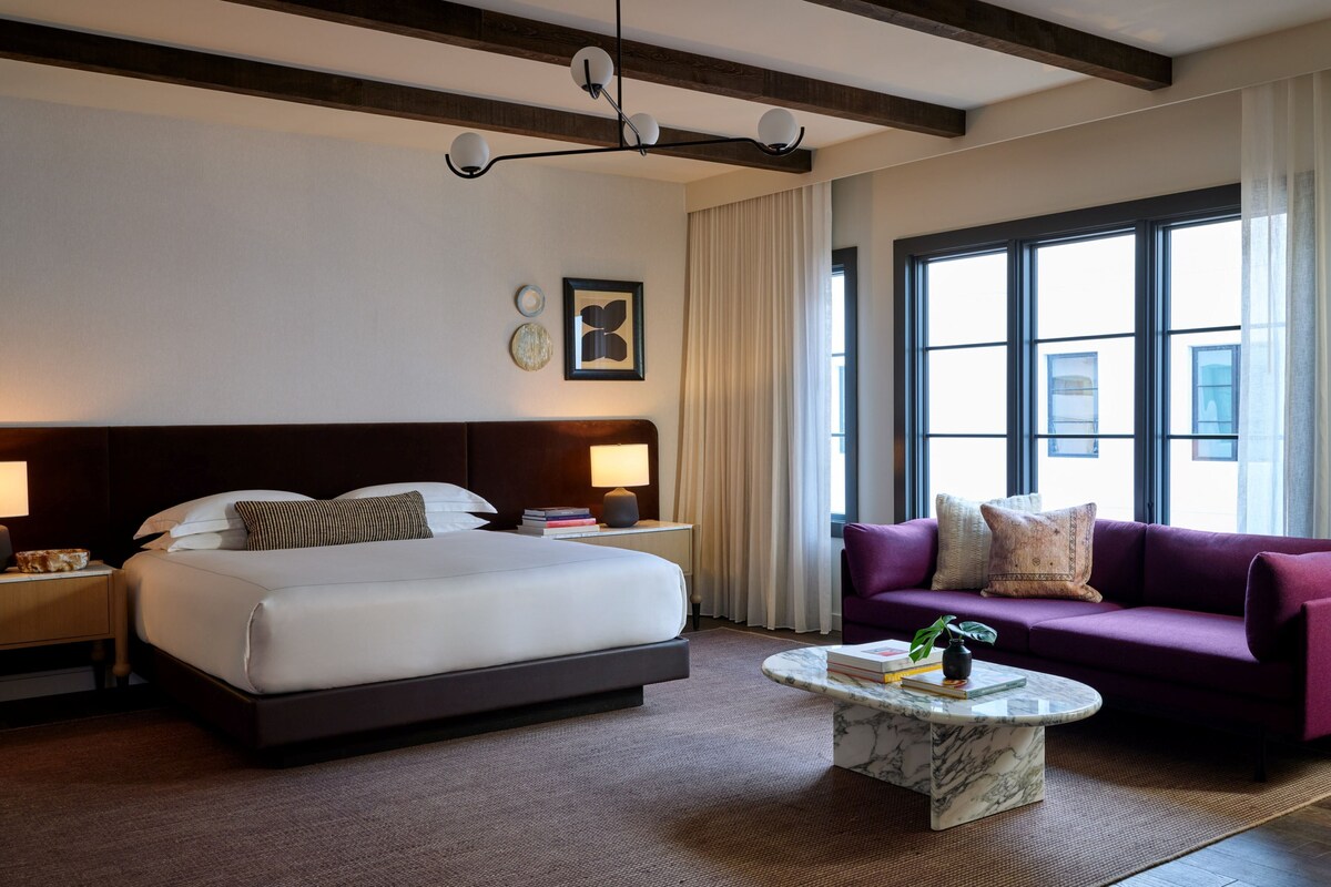 A hotel room with a large bed, a purple sofa, a marble coffee table, and a modern chandelier.