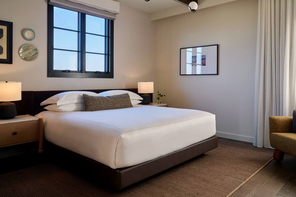 A modern hotel room with a large bed, two bedside tables, a window with blinds, a ceiling light fixture, and a framed picture on the wall.