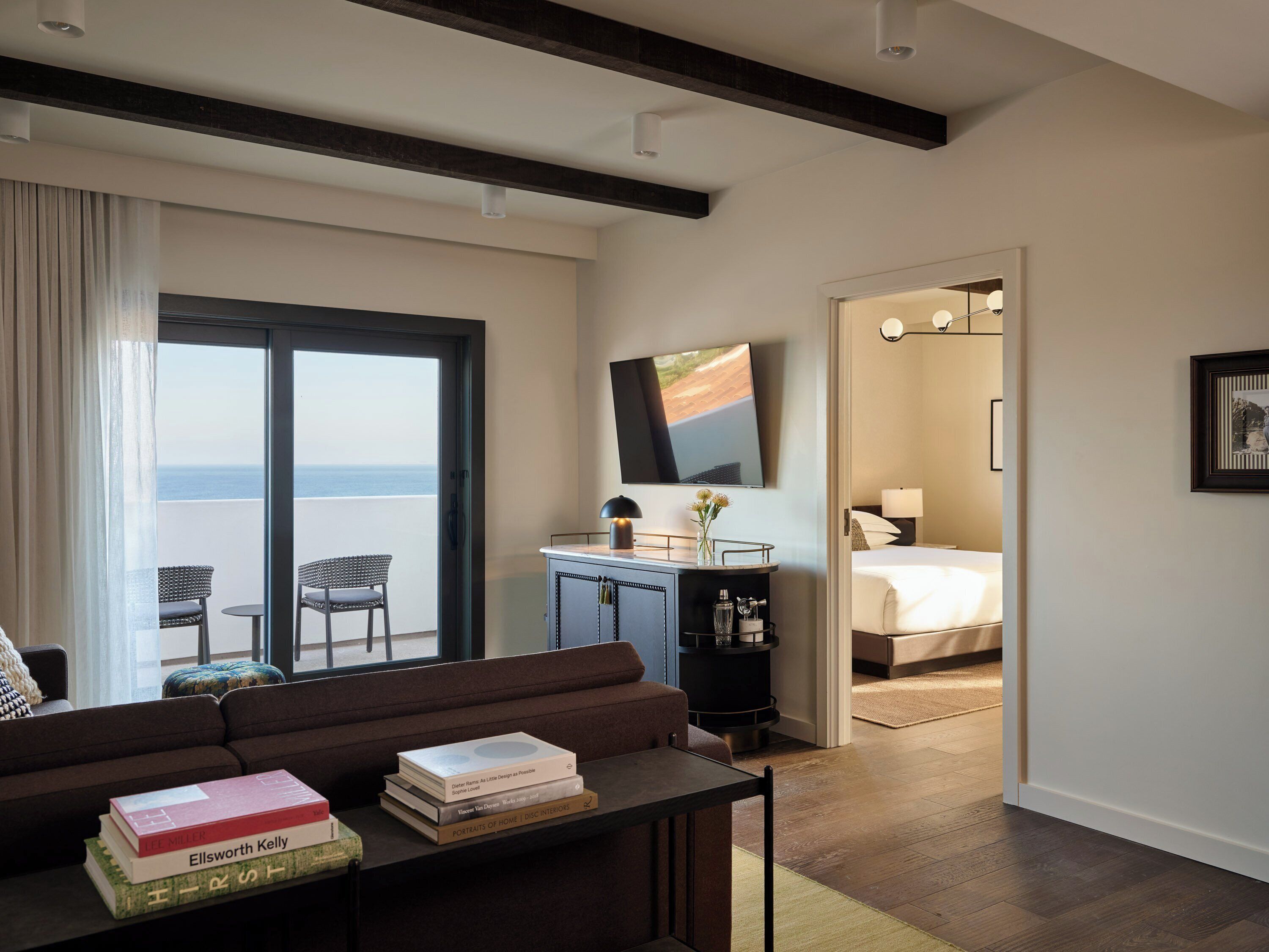 A modern living room with a sofa, a coffee table with books, a desk with a lamp, and a large window overlooking the sea.