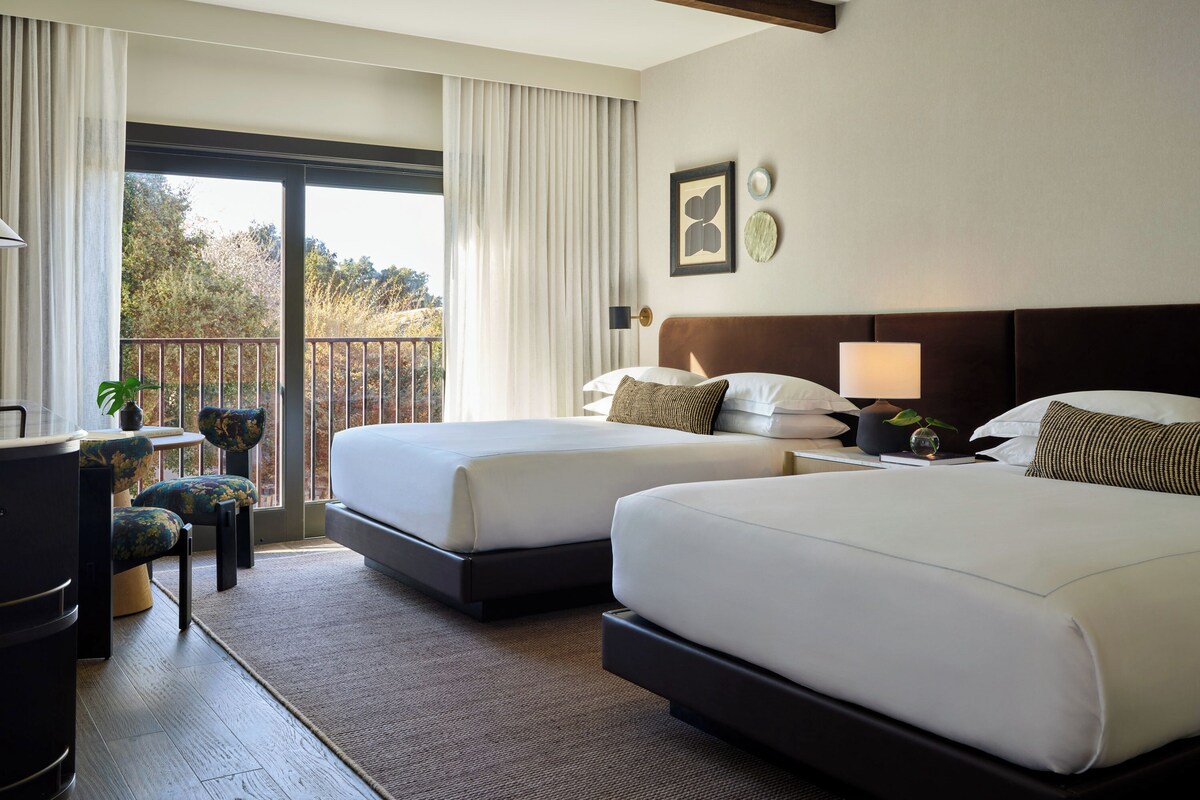 A hotel room with two beds, a desk, a chair, and a balcony with a view of trees.