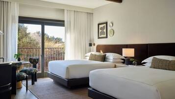 A hotel room with two beds, a desk, a chair, and a balcony with a view of trees.