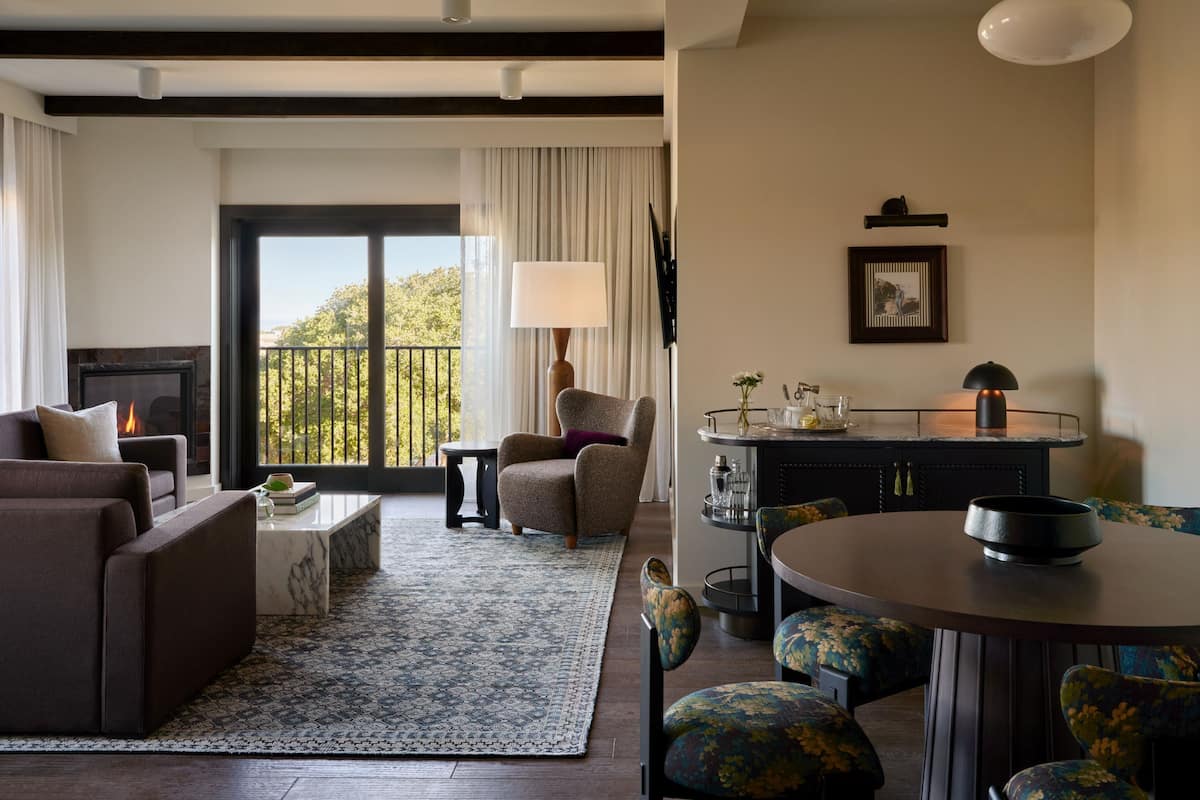 A modern living room with a fireplace, a dining area with a round table and floral chairs, and a view of the outdoors through large windows.