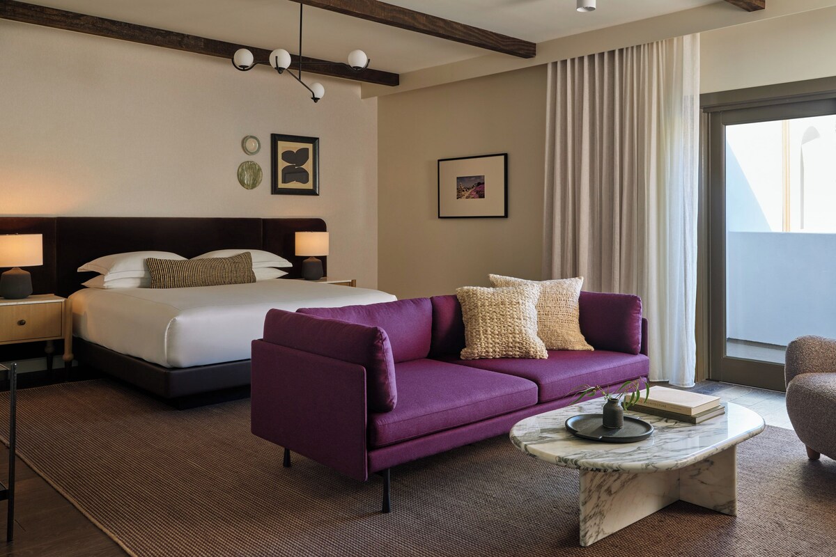 A hotel room with a bed, a purple sofa, a marble coffee table, and a large window with curtains.