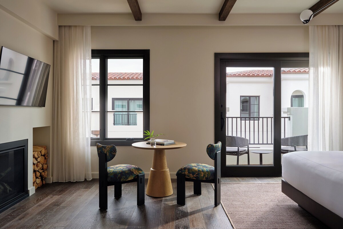 A hotel room with a bed, a TV, a fireplace, a small table with chairs, and a balcony with a view of buildings.